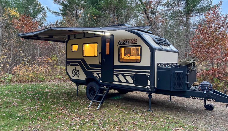 An Outback Series off-road trailer from Imperial Outdoors sits on grass by autumn trees, its awning extended. Warm lights glow inside, a ladder leads to the door, and camping gear is neatly stored at the front.