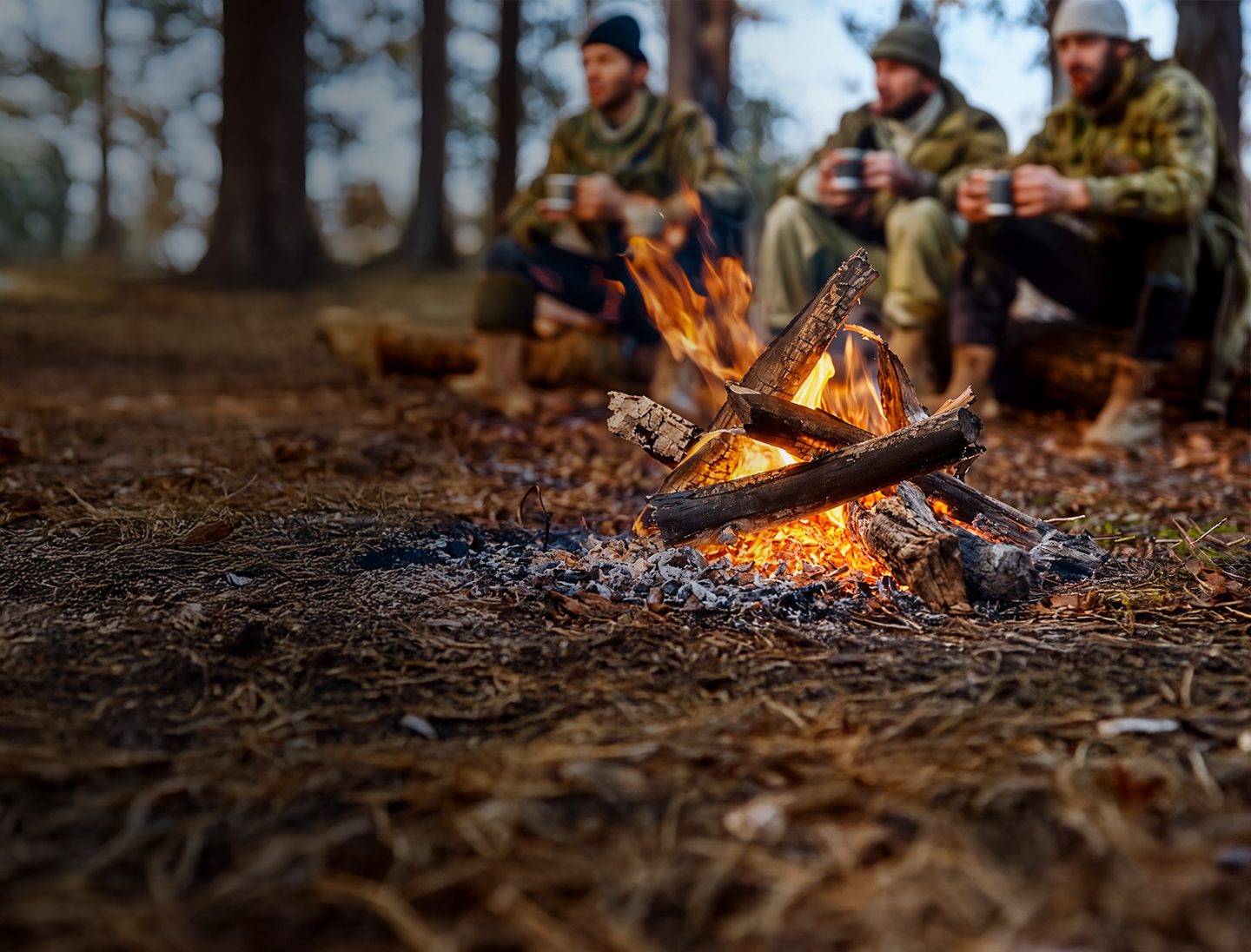 A close-up of a campfire burning with three people in the background, sitting on logs and holding mugs, surrounded by trees—a classic forest scene perfect for adventurers traveling with off-grid all-terrain RV trailers.
