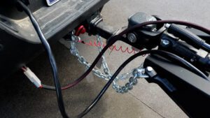 Close-up of a vehicle hitch with safety chains, brake cable, and electrical wiring connected to an Xplore Series off-grid camper trailer on a paved surface.