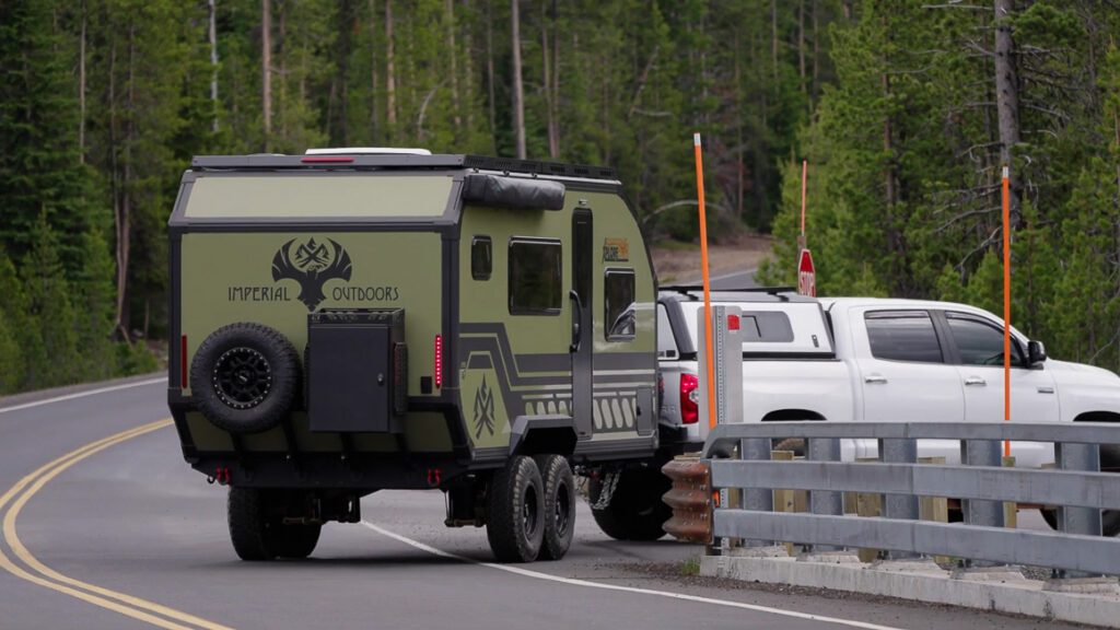 A white pickup truck towing a green Imperial Outdoors Outback Series camper trailer drives on a forested road, crossing a bridge with metal railings and surrounded by trees.