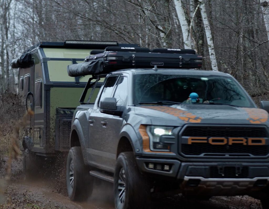 A gray Ford pickup with off-road gear and a rooftop cargo box tows an Imperial Outdoors Sport Series off-road trailer through a muddy forest trail surrounded by bare trees.