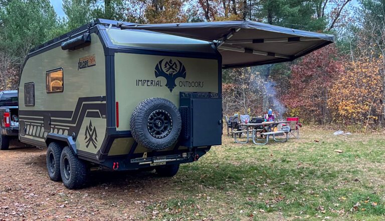 A rugged camper trailer with an open awning is parked in a wooded campsite. A picnic table with people, camping chairs, and a small campfire are visible nearby among autumn trees.