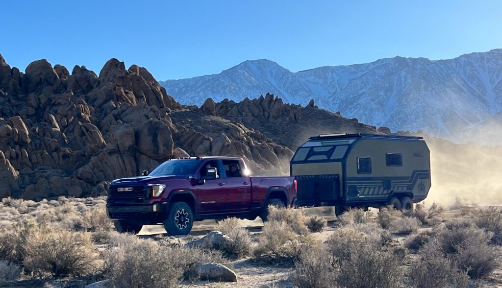 A red pickup truck towing a green camper trailer drives through a dry, rocky landscape with sparse bushes; mountains with snow-capped peaks are visible in the background under a clear blue sky.