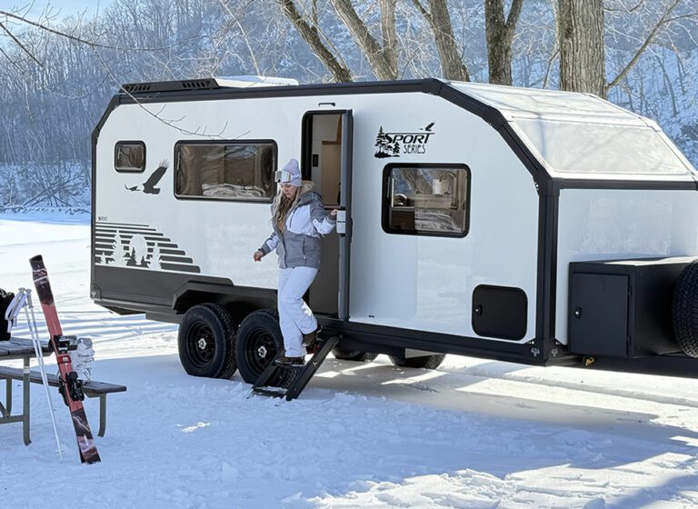 A person wearing winter clothing steps out of a white travel trailer parked on snow, embracing a self-sufficient life. Nearby, skis and poles rest against a picnic table, with snowy trees in the background.
