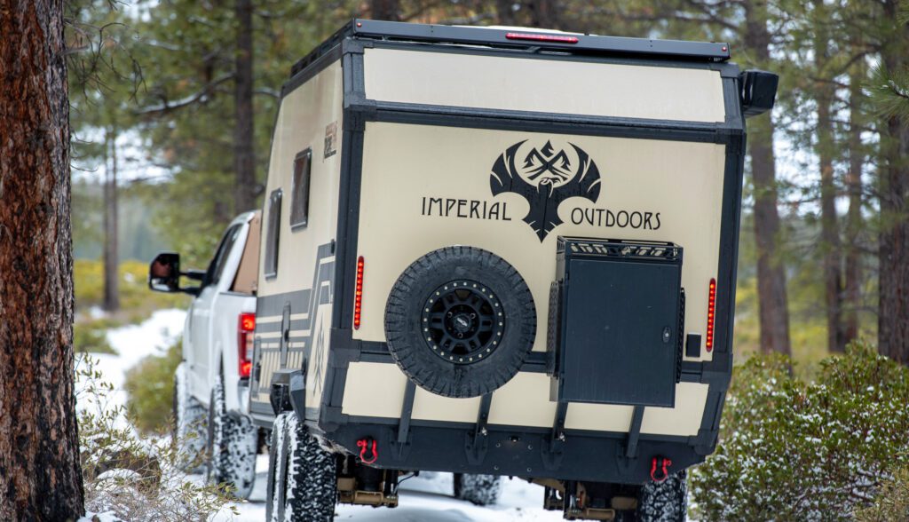 A white pickup truck tows an off-road camper trailer labeled IMPERIAL OUTDOORS through a snowy, forested area with pine trees and shrubs.