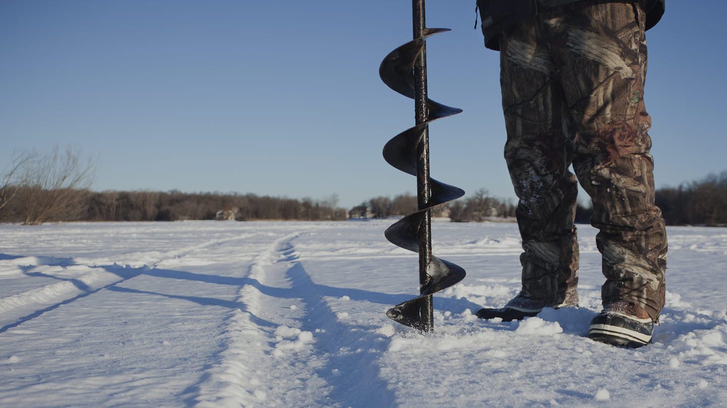 A person in camouflage pants and boots stands on snow, holding an ice auger vertically on a frozen lake, with tire tracks and trees visible in the background under a clear blue sky.