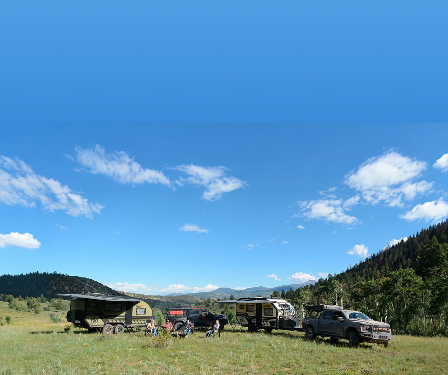 A group of people sits in camping chairs beside a premium off-road adventure trailer, an RV, and a pickup truck on a grassy field with distant mountains, trees, and a bright blue sky with scattered clouds.