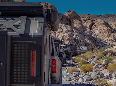 A convoy of off-road vehicles drives through a rocky desert canyon, surrounded by rugged terrain and sparse vegetation under a clear blue sky—no reservations required for this adventure.