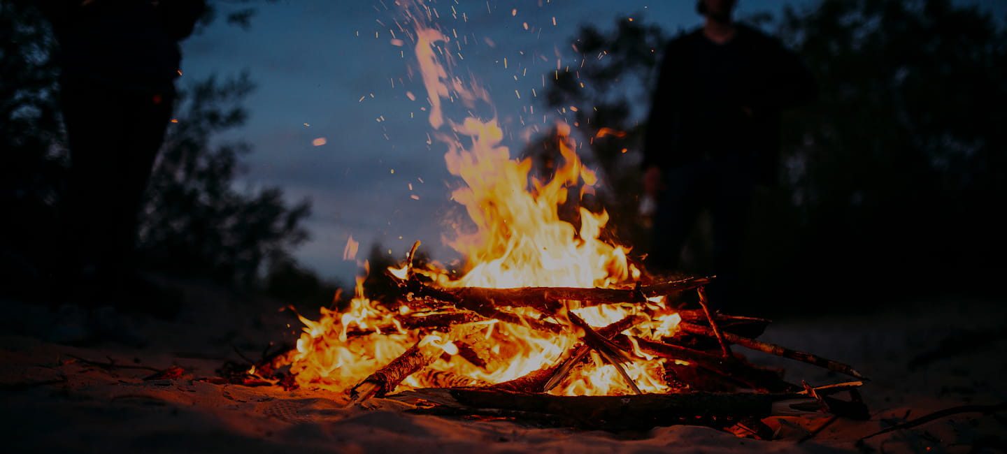 A campfire burns brightly at night, with flames and sparks rising from a pile of wooden logs—perfect for sharing four-season camping tips on your off-grid adventure camping blog. Two people stand in the dim evening light among trees.