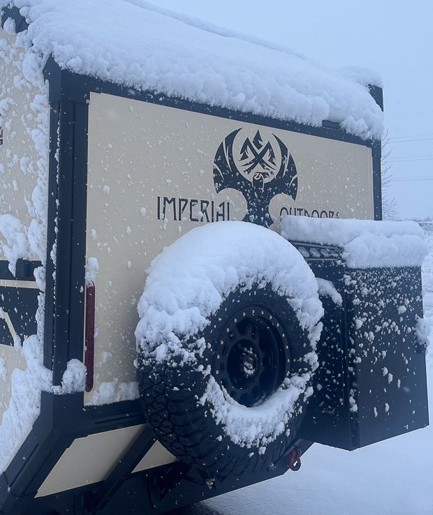 An Imperial Outdoors trailer, built as a four-season off-road camper, sits covered in snow. Drifts pile on the roof, spare tire, and storage box at the back, highlighting its readiness for rugged winter adventures.