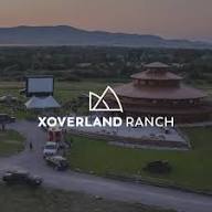 Aerial view of a ranch at sunset with a round building, outdoor movie screen, vehicles, and a logo overlay reading XOVERLAND RANCH with a stylized mountain graphic above the text—capturing The Big Thing happening on the ranch.