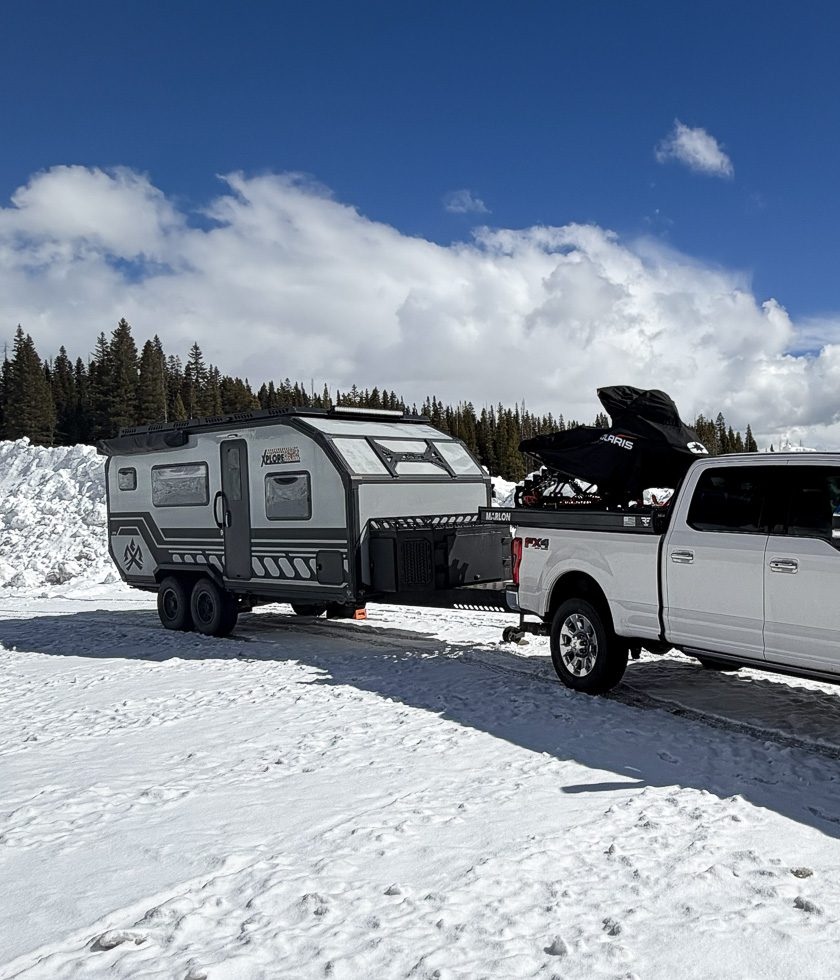 A white pickup truck towing a gray X195 expedition trailer is parked on a snowy landscape with tall snowbanks and pine trees under a partly cloudy sky.