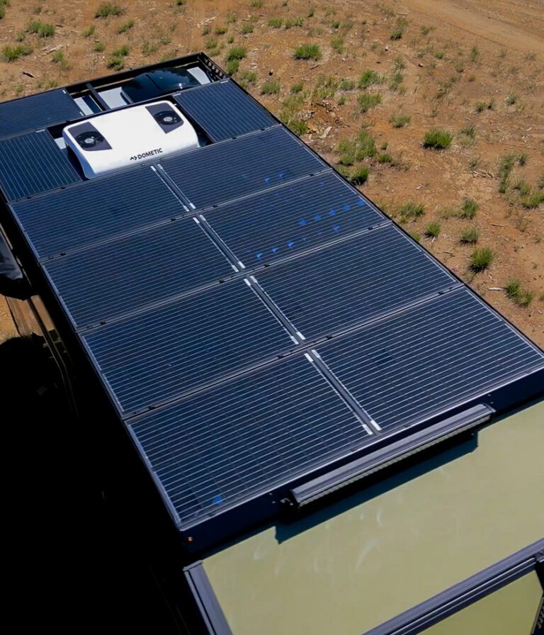A rooftop view of an off-grid camper RV fitted with multiple solar panels, parked on dry, grassy terrain with patches of dirt visible beside the vehicle.