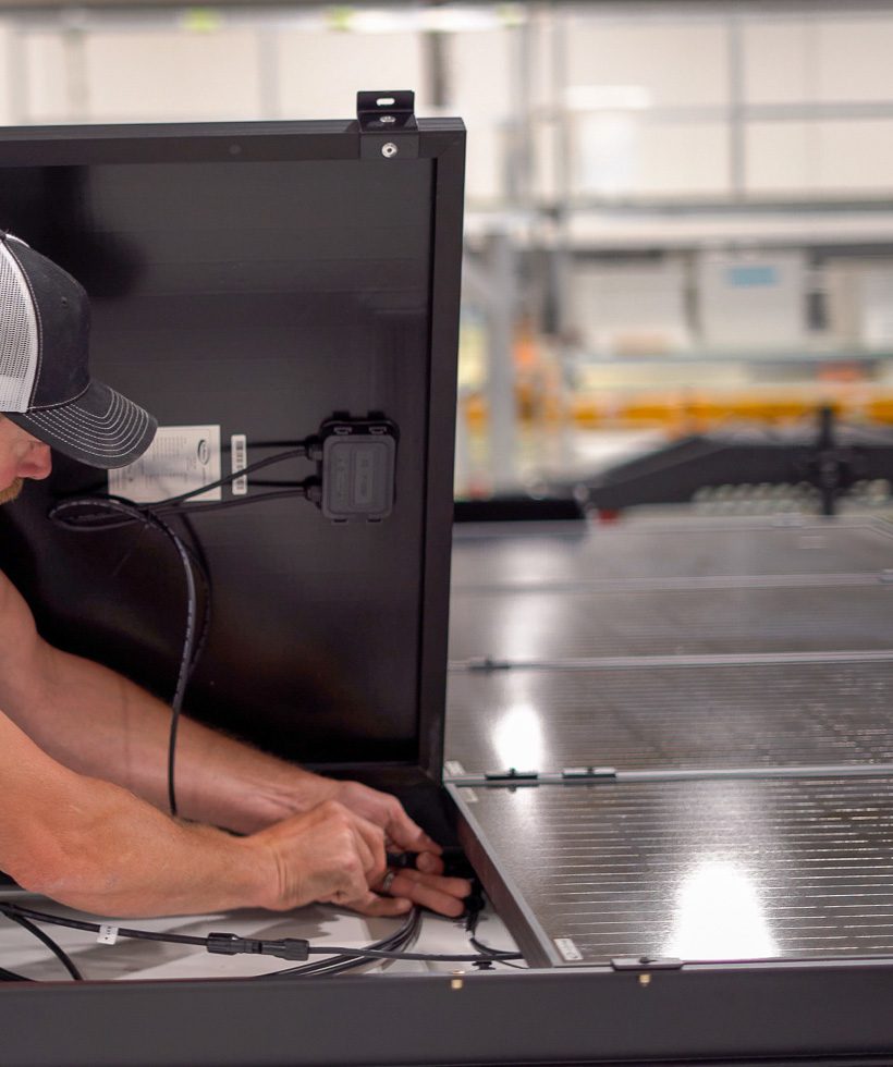 A person wearing a cap is connecting wires to the back of a solar panel in an indoor facility with rows of solar panels visible in the background.
