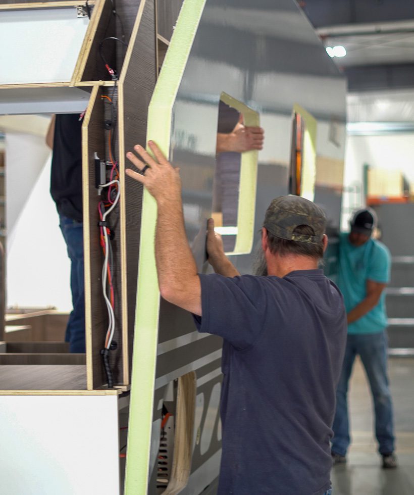 A man wearing a baseball cap is installing foam insulation panels on the side of a partially assembled RV or trailer inside a workshop; another person works in the background.