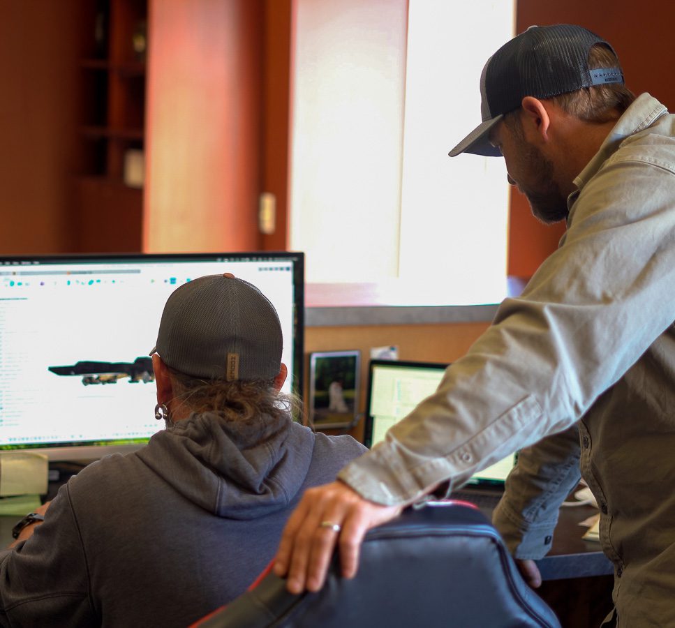 Two men wearing caps work together at a computer desk; one is seated using the computer while the other stands beside him, leaning with one hand on the chair and looking at the monitor.
