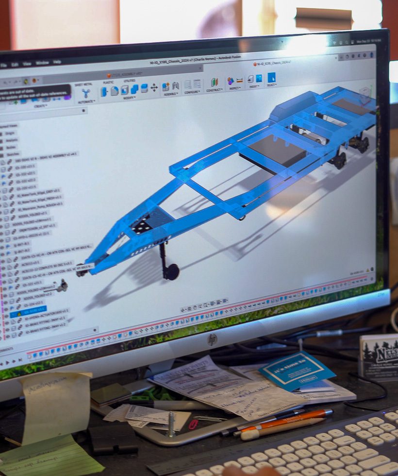 A computer monitor displays a 3D CAD model of a blue trailer frame. The workspace around the monitor is cluttered with papers, pens, a notebook, business cards, and sticky notes.