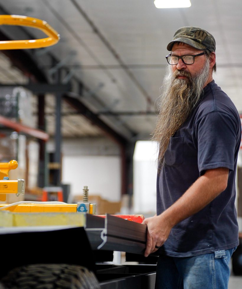 A man with a long beard, glasses, and a camouflage cap works in a warehouse, holding a rectangular object while standing near industrial equipment and shelves.