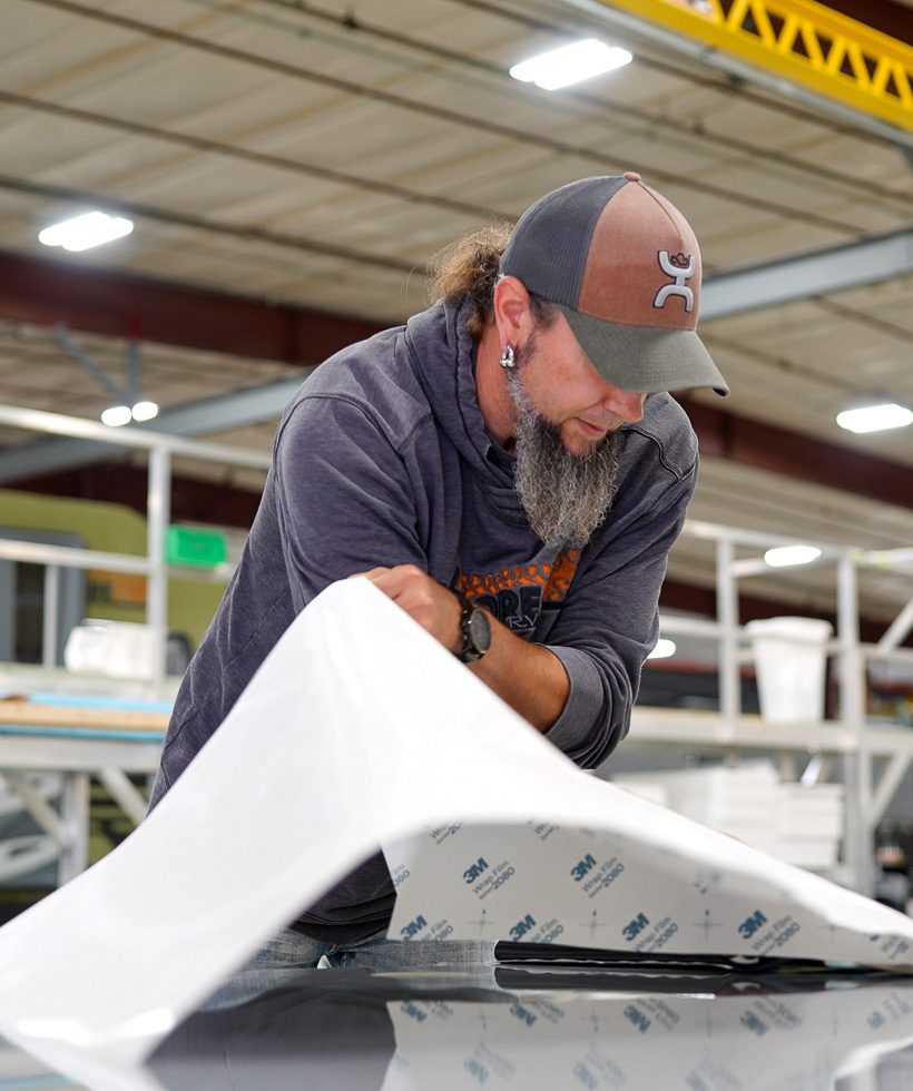 A man with a gray beard in a baseball cap and hoodie carefully applies a large sheet of adhesive material onto a flat surface in an industrial workshop setting. Shelves and tools are visible in the background.