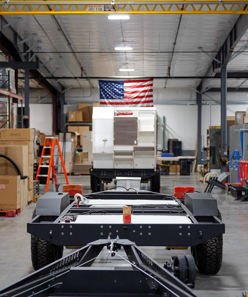 A factory or warehouse interior with a large American flag on the back wall, machinery and boxes around, and a vehicle chassis in the foreground facing another partially assembled vehicle.