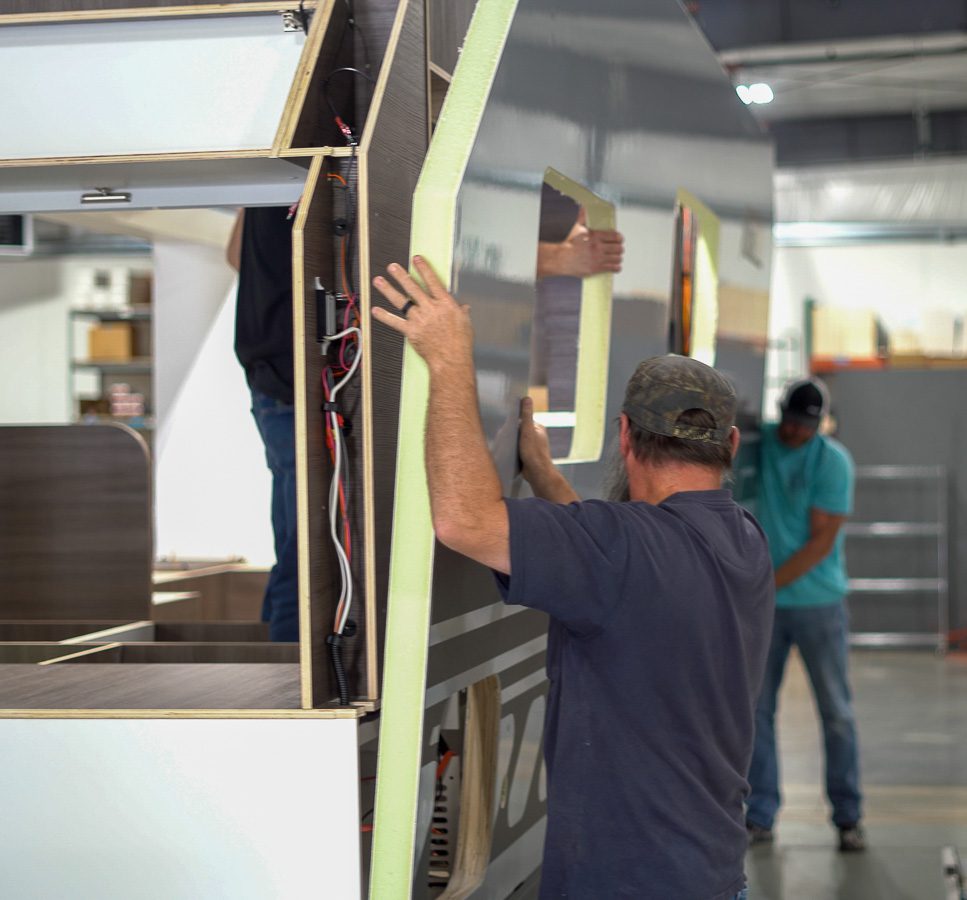 A man wearing a cap installs a wall panel with a cutout window onto a partially constructed trailer indoors; another worker and construction tools are visible in the background.