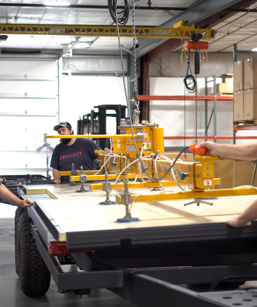 A worker in a cap operates a yellow industrial vacuum lifter to move a large flat panel onto a trailer bed inside a warehouse, while another person assists from the side.
