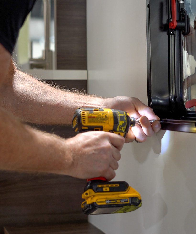 A person uses a yellow cordless power drill to fasten a screw into the frame of a window set in a white wall, with part of their arms and hands visible.