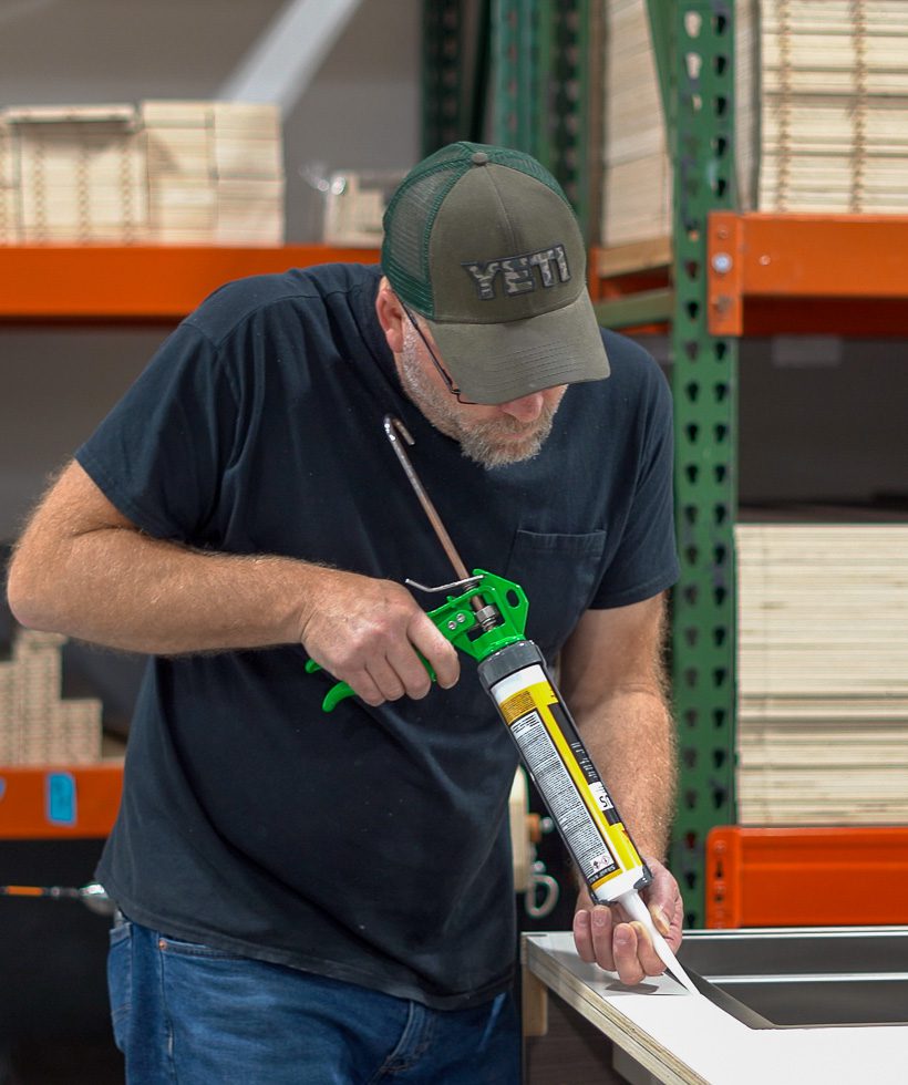 A man wearing a green cap and black t-shirt applies sealant with a caulking gun to the edge of a white surface in a workshop with shelves in the background.