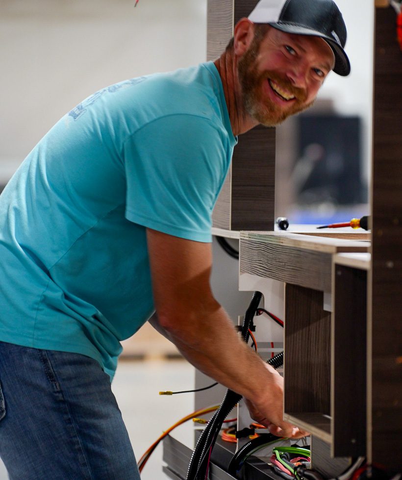 A smiling man in a blue shirt and black cap works on electrical wiring inside a wooden structure, possibly assembling or repairing equipment.