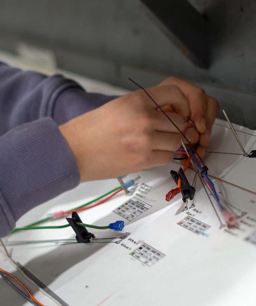 A person’s hands work with colored wires and small clamps on a white surface covered with diagrams and labels, possibly assembling or organizing electrical components.