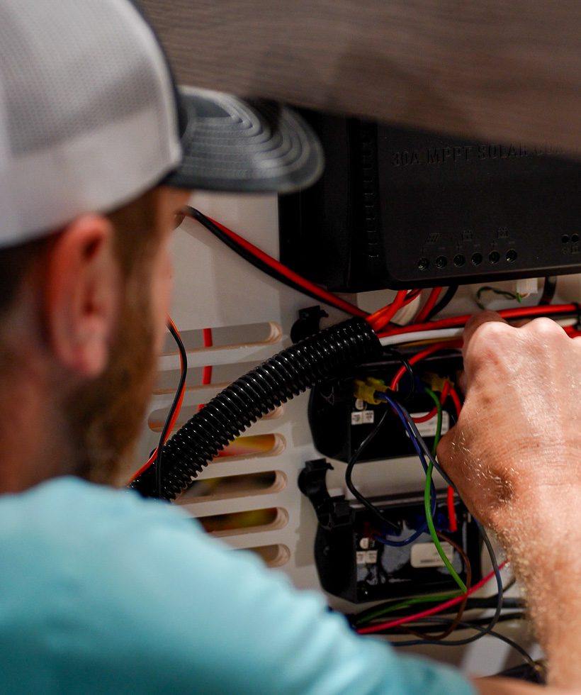 A person wearing a cap works on electrical wiring inside a panel, connecting various colored wires and components. The focus is on the hands and wires, with the persons face partially out of view.