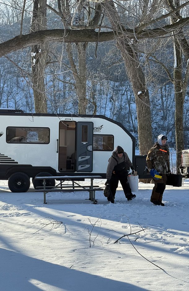 Two people in winter clothing stand near a picnic table in the snow beside a white Imperial Outdoors trailer, showcasing the comfort of four-season off-road campers amid bare trees and a snowy landscape.