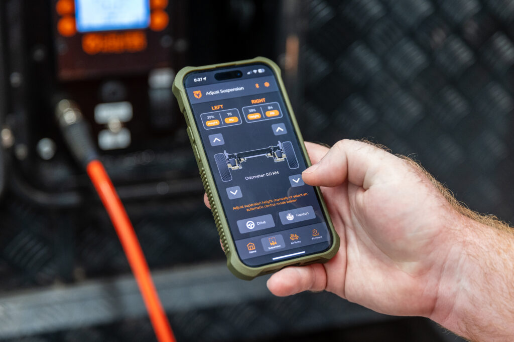 A hand holds a smartphone displaying a vehicle suspension control app, showing options to adjust front and rear suspension settings. A cable and electronic device are visible in the blurred background.