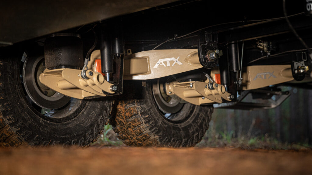 Close-up of off-road vehicle suspension system with large, mud-covered tires and beige components labeled ATX, set outdoors on a dirt surface.