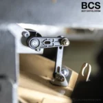 Close-up of a mechanical component with a metal arm and bolts, part of a body control system. The BCS Body Control System logo is visible in the top right corner.