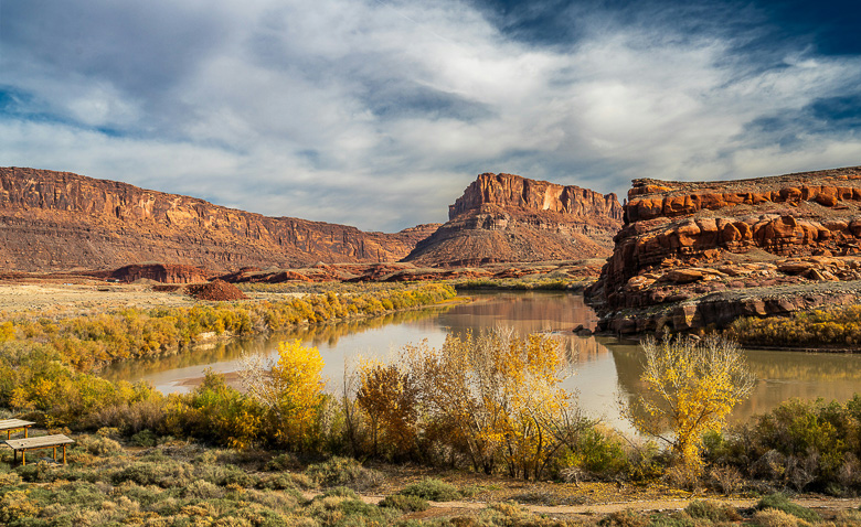 A scenic view of a river winding through a desert canyon with red rock cliffs, yellow autumn trees, and a dramatic cloudy sky in the background—an ideal spot for off-grid fall camping and those seeking unique shoulder season camping experiences.