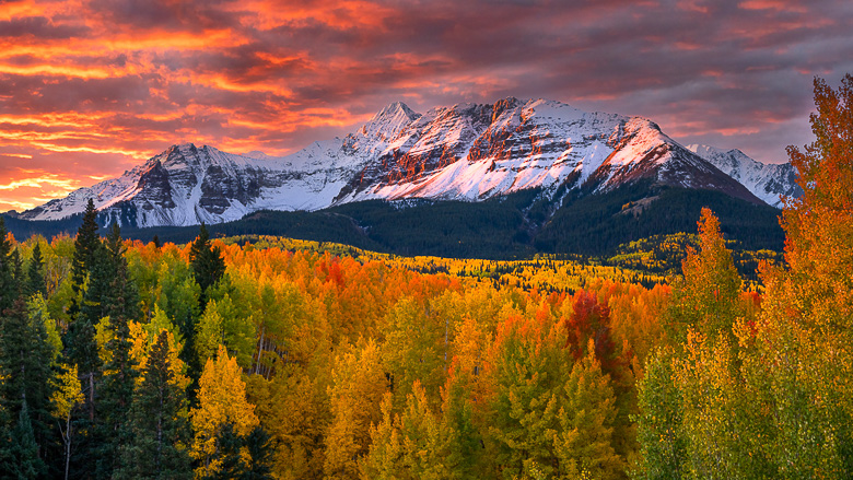 Snow-capped mountains under a vibrant orange sunset sky, with a forest of green, yellow, and orange trees in the foreground—an idyllic scene perfect for off-grid fall camping and exploring top fall camping destinations.