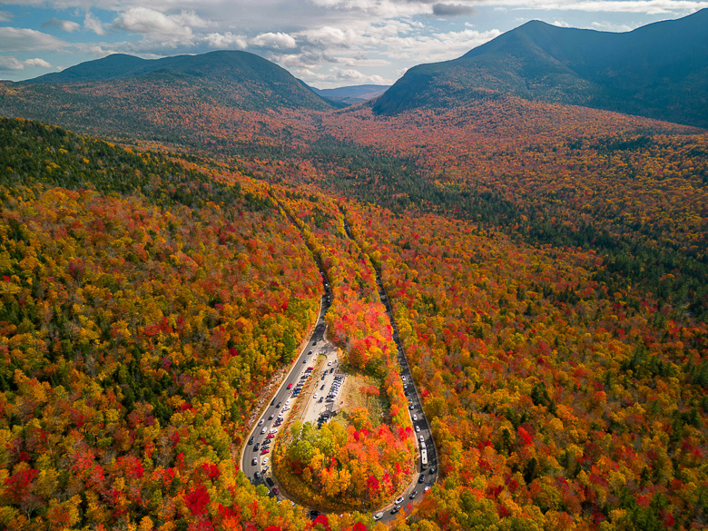 Aerial view of a forest with vibrant fall foliage, a curving road lined with parked cars, and mountains in the background under a partly cloudy sky—an idyllic spot perfect for those seeking off-grid fall camping adventures.