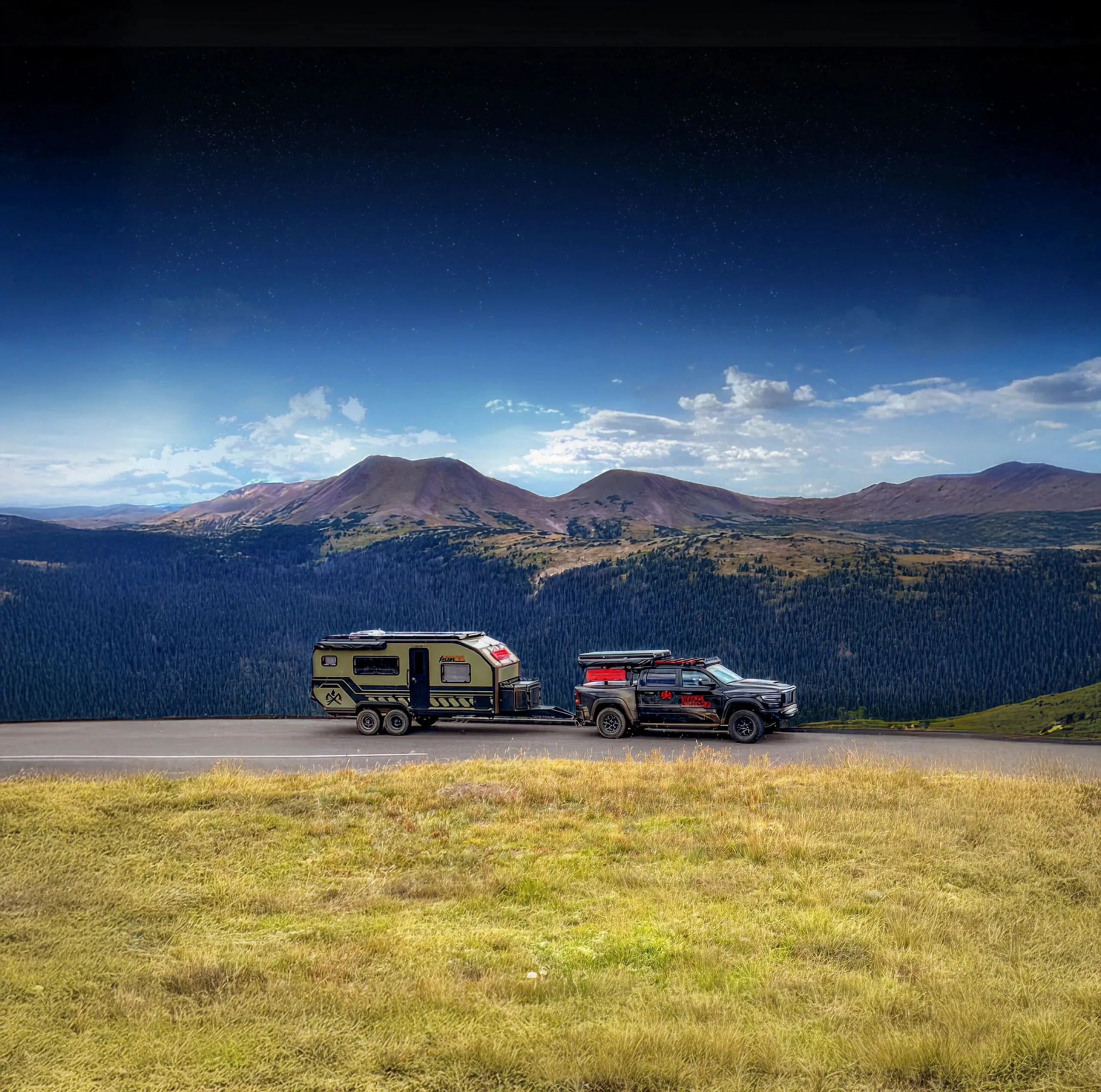 A truck towing an Imperial Outdoors Xplore Series camper trailer drives on a road through a grassy area with mountains and forests in the background under a bright blue sky with scattered clouds.
